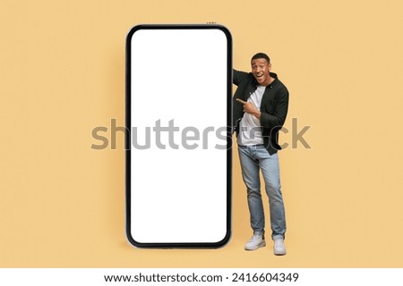 Nice mobile app. Excited handsome young black man standing by huge cell phone with white empty screen, showing great online offer, isolated on beige background, mockup, copy space