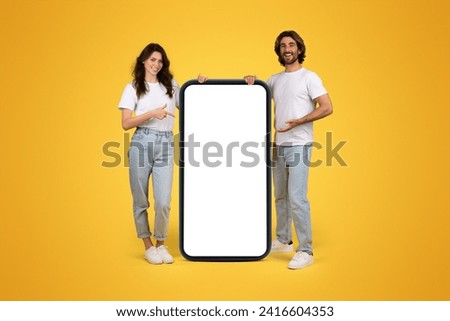 Smiling young european guy and woman in casual point fingers at huge smartphone with empty screen, isolated on orange background studio, collage. Website, app, blog presentation, recommendation