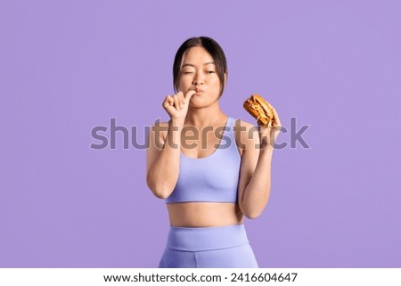 Fit asian woman in workout clothing, looking at tasty burger in her hand and licking her finger, contemplating diet choices against purple backdrop Royalty-Free Stock Photo #2416604647