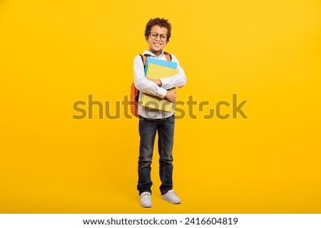 Smart-looking schoolboy with glasses hugging school folders and wearing backpack, standing confidently against cheerful yellow background, full length Royalty-Free Stock Photo #2416604819