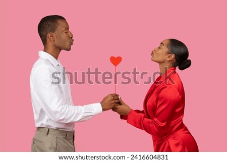Side profile of black man in white shirt and woman in red dress romantically holding heart on stick together, symbolizing shared love against pink backdrop Royalty-Free Stock Photo #2416604831