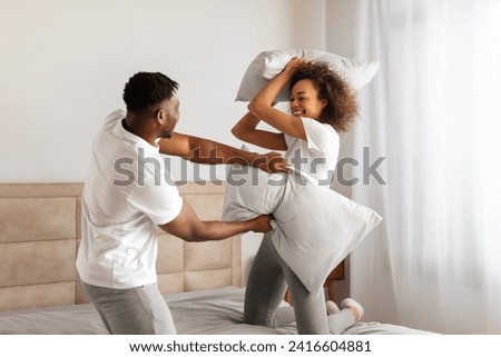 Playful loving black couple enjoys carefree pillow fight, filling cozy bedroom with laughter as they having fun and fighting with pillows in modern home interior in the morning Royalty-Free Stock Photo #2416604881