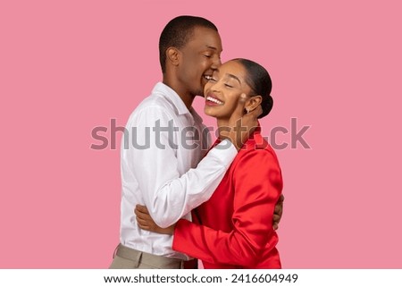 Joyful black man in white shirt affectionately kissing cheek of smiling woman in red dress, sharing tender moment against pink background Royalty-Free Stock Photo #2416604949