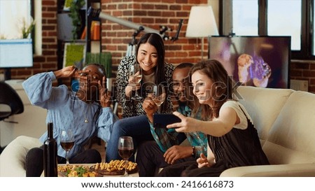BIPOC friends taking group pictures with selfie camera smartphone at apartment party. Joyful mates in modern home posing for photo with wine and champagne glasses, making gestures