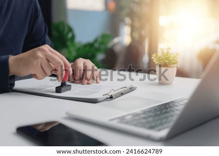 Individual in an office setting applies a stamp to a document contract legal agreement, signifying approval or verification certification permit visa next to an open laptop.
