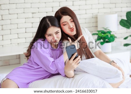 Two Asian cheerful happy female girlfriends in casual outfits sitting resting relaxing holding touchscreen smartphone showing two finger sign smiling taking selfie photo together on bed in bedroom.
