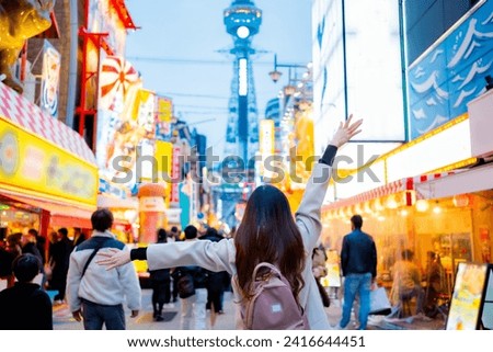 Asian female tourist Traveling and having fun. And she was taking photo with a cell phone camera at Night street with many restaurant around Tsutenkaku Tower in Shinsekai district of Osaka, Japan.