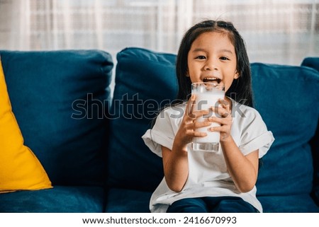In a cozy apartment a cheerful Asian girl drinks milk from a glass while sitting on the sofa. This heartwarming picture emphasizes daily health care and the joyful innocence of childhood.