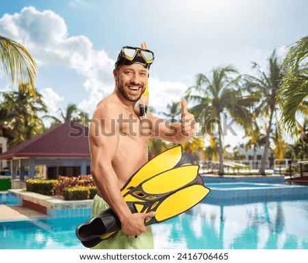 Cheerful young man in swimwear with a diving mask and snorkeling fins gesturing thumbs up by a swimming pool