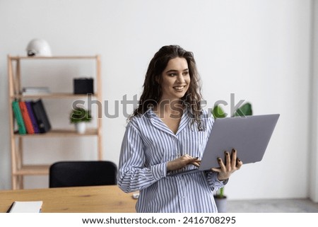 A young smiling business woman is standing in the office by the window with a laptop in her hands. Woman manager in glasses and white shirt