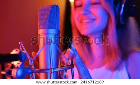 Young woman records a song into a studio microphone under neon LED lighting, blue and purple, in a recording studio
