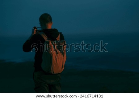 back rear view of one alone backpacker Man with rucksack, cell phone standing on peck of rock, taking photos of the view. Man on top of a mountain photographing sky above Black sea or pacific ocean.