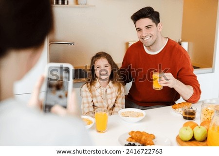 Portrait smiling young family, having breakfast with little daughter, drinking orange juice, mother holding mobile phone taking pictures, in cozy room, at home. Concept of parenthood, food, breakfast