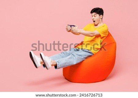 Full body young man he wearing yellow t-shirt casual clothes sit in bag chair hold in hand play pc game with joystick console isolated on plain pastel light pink background studio. Lifestyle concept