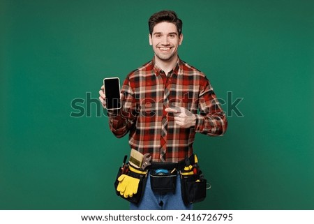 Young employee laborer man wear red shirt use point on blank screen workspace area mobile cell phone isolated on plain green background. Instruments for renovation apartment room. Repair home concept