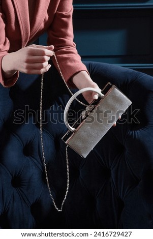 Silver metallic sparkling purse held by woman hands in pink suit on a blue quilted velour armchair background. Creative shiny woman hand bag fashion studio photography. Royalty-Free Stock Photo #2416729427