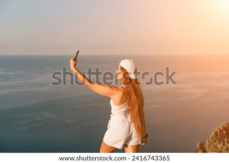 Selfie woman sea. The picture depicts a woman in a cap and tank top, taking a selfie shot with her mobile phone, showcasing her happy and carefree vacation mood against the beautiful sea background