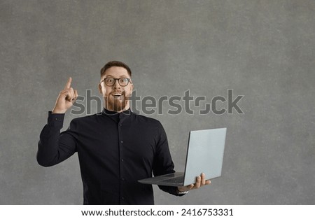 Portrait of funny nerdy bearded young man in glasses isolated on grey background holding laptop computer, looking at camera, smiling and pointing index finger up surprised by a good and smart idea