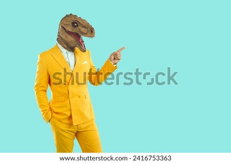 Stylish man in funny dinosaur mask, points to an empty space to copy. Funny showman in bright yellow suit and white shirt on an isolated pale blue background.