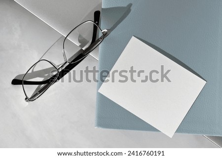 Empty paper card, light blue notebook, eyeglasses and laptop on marble gray desk background, business card template, flat lay, top view.