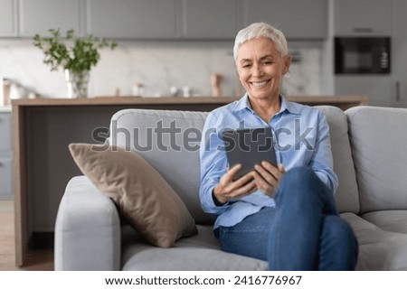Happy senior woman sitting with digital tablet, enjoying gadget use and online world, websurfing or watching movie online on computer, relaxing on sofa at home living room Royalty-Free Stock Photo #2416776967