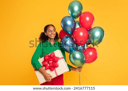 Birthday surprise. Cheerful black teenager girl holds big wrapped gift box with bow and bunch of colorful balloons standing on yellow studio backdrop, smiling to camera, celebrating bday holiday