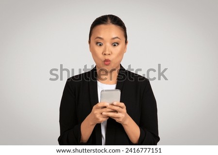 Surprised Asian businesswoman looking at smartphone with an exaggerated expression of shock, possibly after receiving unexpected news or a message, isolated on gray background, studio Royalty-Free Stock Photo #2416777151