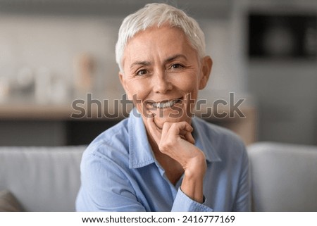Aged Beauty. Portrait of relaxed gray mature lady with short haircut sitting on the couch indoor in living room and posing. Positive older woman looking and smiling at the camera Royalty-Free Stock Photo #2416777169