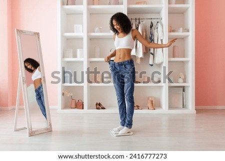 Curly-haired latin woman proudly showing her weight loss success by wearing too-large jeans, reflected in a full-length mirror in a light room Royalty-Free Stock Photo #2416777273