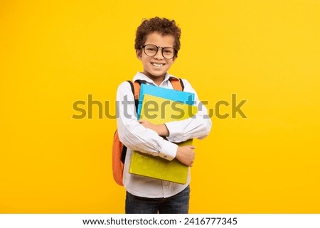 Smiling nerdy boy with curly hair and glasses, clutching school books and wearing an orange backpack, ready for class against yellow backdrop