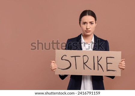 Upset young woman holding cardboard banner with word Strike on brown background, space for text