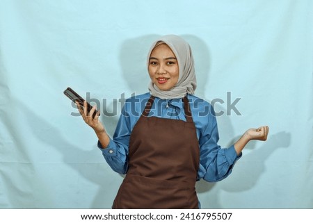 young Asian muslim woman wearing hijab and brown apron holding mobile phone while show winner gesture isolated and smiling showing her teeth  over white background.housewife muslim lifestyle concept