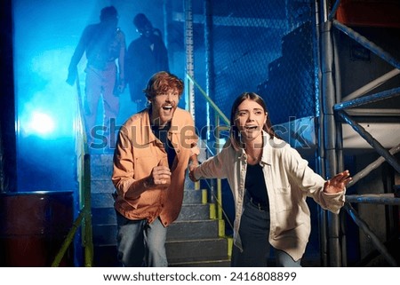 scared man and woman running from freaky people in gas masks and orange ppe suits in escape room Royalty-Free Stock Photo #2416808899