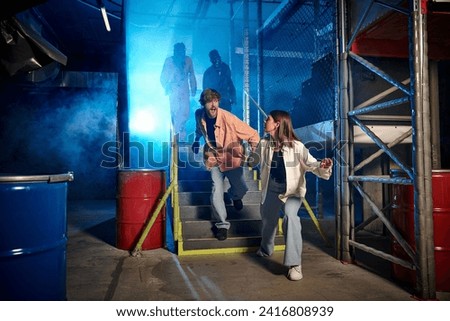 young man and woman running from scary people in gas masks and orange ppe suits in escape room Royalty-Free Stock Photo #2416808939