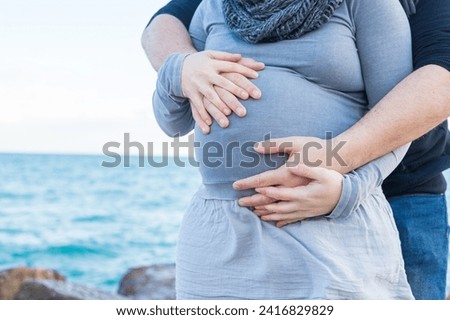 Hands clasped on pregnant belly on the beach. A pair of intertwined hands hug and caress a pregnant belly. One pair of affectionate hands are above the belly and the other pair is below. Royalty-Free Stock Photo #2416829829