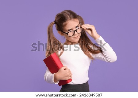 Cute schoolgirl with book on violet background Royalty-Free Stock Photo #2416849587
