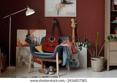 Soft comfortable armchair with acoustic guitar and pile of clothes standing by wall with paintings, lamp, green plant and electric string instrument