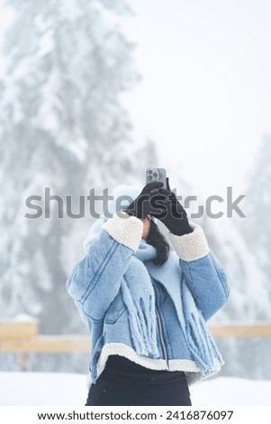 Woman wearing winter clothes taking a photo with mobile phone on a snowy day outside around pine trees 