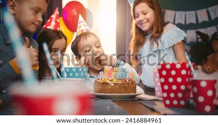 Cute small Caucasian boy leaning on the table and looking at the cake at his birthday party among friends takes a piece of celebration cake and then bite it. Cute boy eats cake on his birthday.