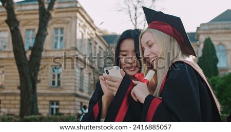 Close up of Caucasian pretty young blond girl in academic cap and gown demonstrating some photos or videos on the smartphone to her Asian female friend