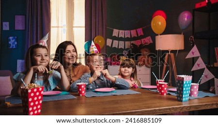 Caucasian cute little boy with his siblings and mother sitting at the table and waiting for the birthday cake with candles while somebody carrying it and putting on the table.
