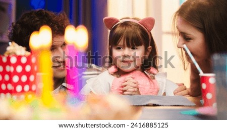 Close up of the small cute girl in funny ears applauding while sitting among happy mother and father in front of the birthday cake with candles.