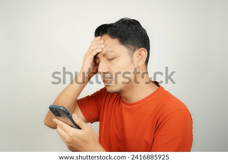 Asian man in orange t-shirt seen holding head holding problems looking at message on smartphone on isolated background