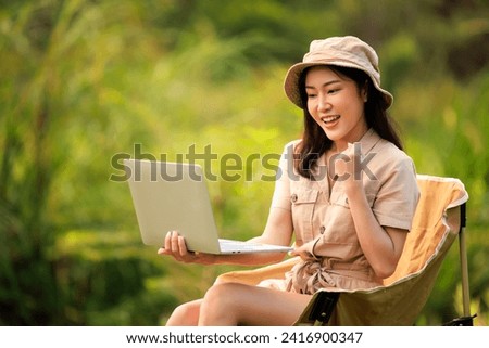Young asian woman in safari dress sitting on chair using laptop in the nature, freelance work adventure and campground travel concept, 