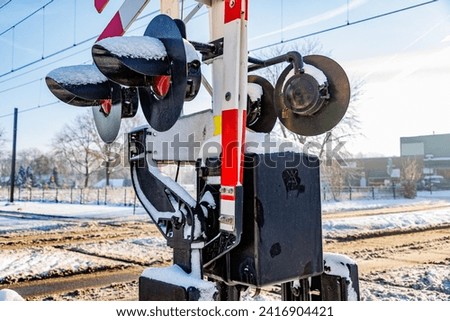 Closeup of warning lights and raised crossing barriers at railway crossing, snow covered train tracks in blurred background, sunny day after heavy snowfall in Beek - Elsloo, South Limburg, Netherlands