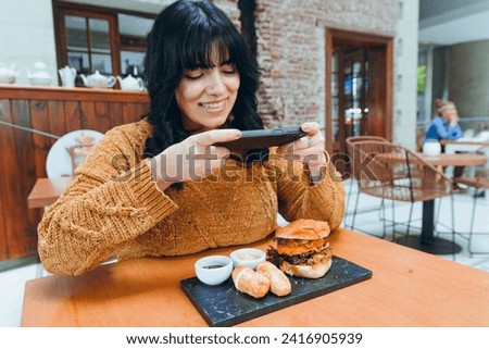 young hungry latin woman sitting in fast food restaurant happy taking photo of hamburger she is eating with her phone, technology and food concept, copy space.