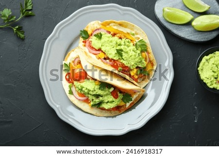 Delicious tacos with guacamole and vegetables served on black table, flat lay