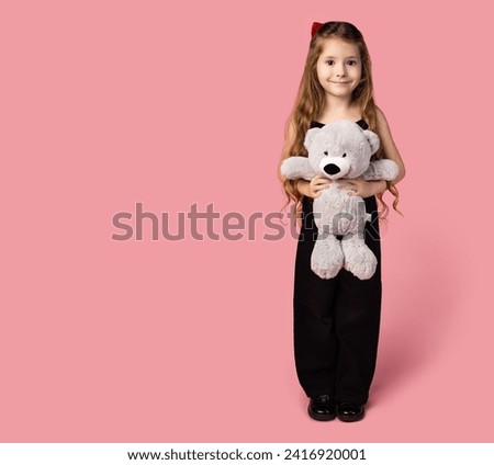 Full-length portrait of a little girl dressed in black denim, with a gray teddy bear in her arms, looking happily at the camera, dedication to the love of toys. Picture on light pink background.