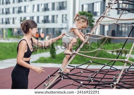 Young mother aids her little daughter as she navigates rope climbing frame at playground with care.