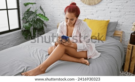 Young beautiful redhead woman, comfortably relaxed in pajamas, smiling while using smartphone on cozy bed, enjoying quiet morning indoors in her homey bedroom